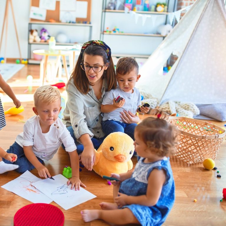 Eine Frau spielt mit Kindern auf dem Boden, umgeben von Spielzeug und Farbstiften.