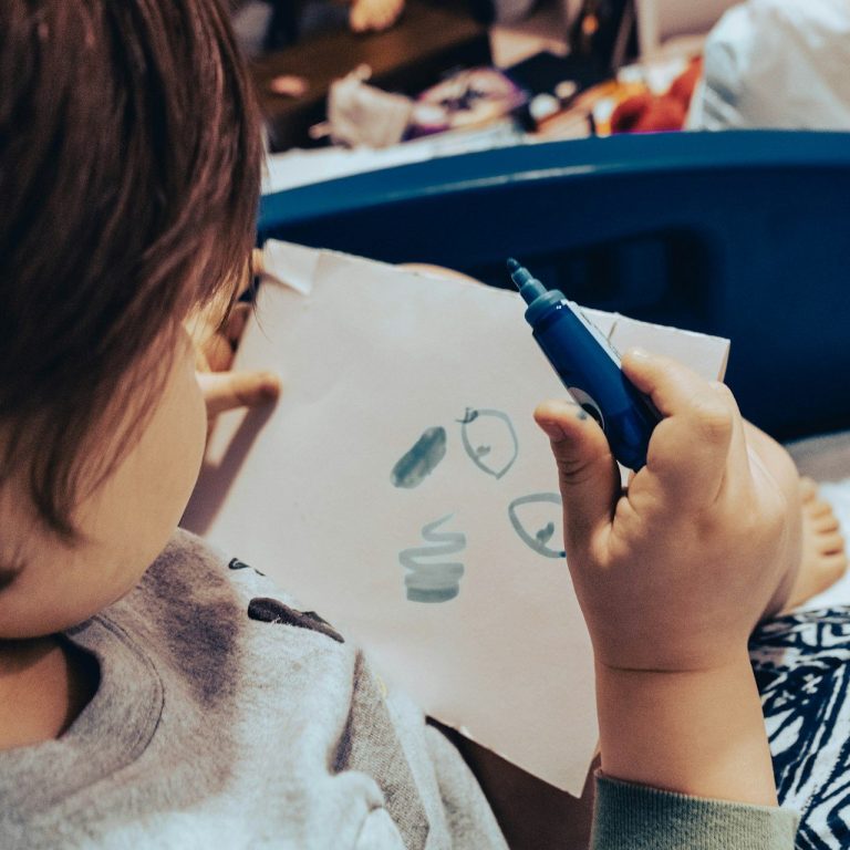 Kind malt mit blauer Farbe auf weißem Papier, umgeben von verschiedenen Gegenständen.