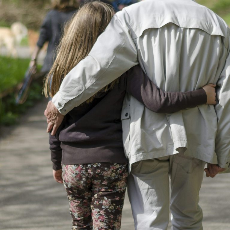 Ein Erwachsener und ein Kind gehen nebeneinander, Arm in Arm, auf einem Gehweg.