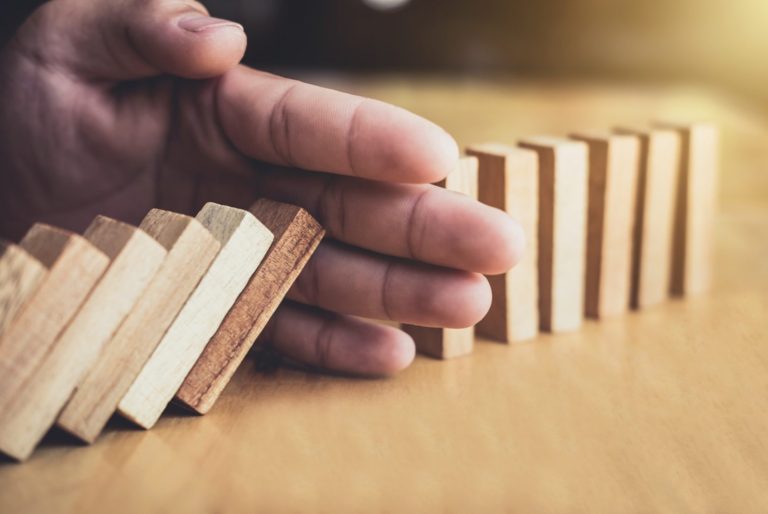 Eine Hand hält Holzdominos zurück, die kurz davor stehen, umzufallen.