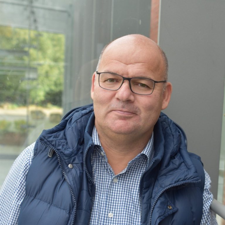 Mann mit Brille und Weste, lächelt freundlich in einem modernen Umfeld.